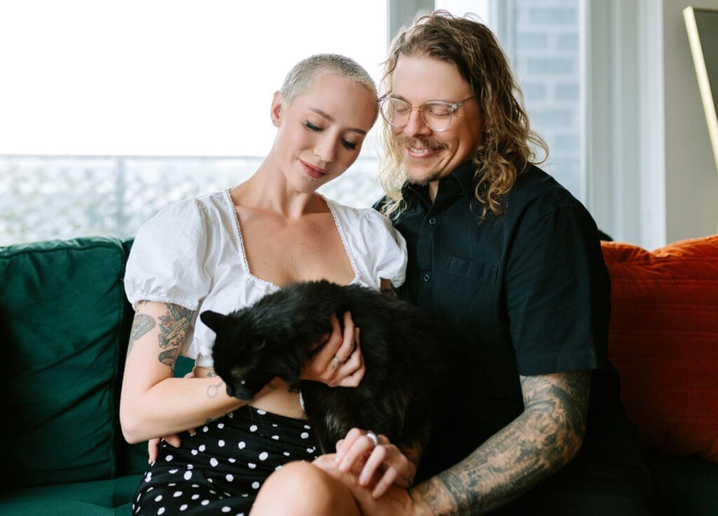 A couple poses at home with their black cat